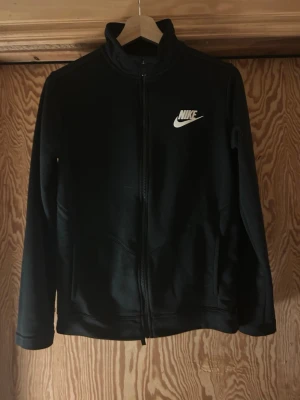Svart zip hoodie från Nike - Säljer en svart zip hoodie från Nike med vit logga på bröstet. Hoodien har hög krage, dragkedja framtill och två fickor. Tillverkad i mjukt material som känns skönt mot huden. Perfekt för en sportig och avslappnad stil.