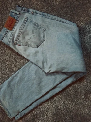 Ljusblå raka jeans från Levi's W34 L32 - Snygga ljusblå jeans från Levi's med klassisk femficksdesign och raka ben. Jeansen har en ikonisk läderpatch bak i midjan och är tillverkade i denim. Perfekta för en avslappnad och trendig stil. Tyvärr en liten slitning vid vänster ben