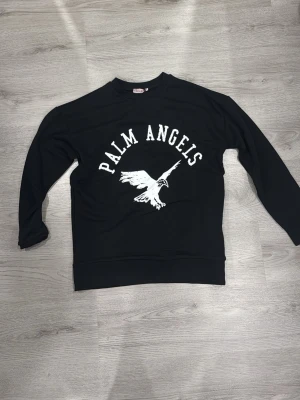 Svart Palm Angels hoodie med tryck - Svart hoodie från Palm Angels med stort vitt tryck av en fågel och texten 'PALM ANGELS' på bröstet. Klassisk passform med rund hals och långa ärmar. Perfekt för dig som gillar streetwear och vill sticka ut med en snygg logga.