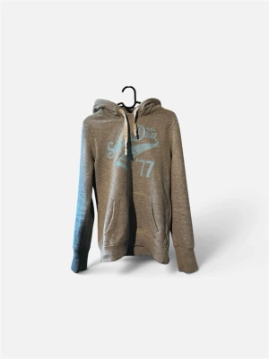 Grå hoodie med tryck och ficka - Snygg grå hoodie med ljusblått tryck framtill och texten 'Super Dry 77'. Klassisk känguruficka, huva med vita snören och ribbade muddar. Perfekt för chill dagar och enkel att matcha med jeans eller joggers.