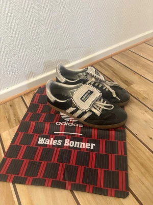 Adidas x Wales Bonner svart sneaker - Unika Adidas x Wales Bonner sneakers i svart mocka med vita detaljer och klassiska tre ränder på sidan. Skorna är i okej skick som man ser lite defekter(märket i innersulan, skosnöret och lädret bak). Skorna har en speciell tunga med dekorativa sömmar och beige snören. Yttersulan är i gummi och designen är retroinspirerad och stilren. Kvitto finns fråga efter de!