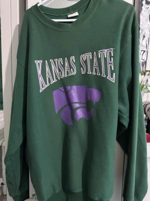 Grön Kansas State collegetröja - Säljer en grön collegetröja med trycket 'Kansas State' i vitt och lila samt ett stort lila logomärke på bröstet. Tröjan har rund halsringning och långa ärmar. Perfekt för dig som gillar college-stil.