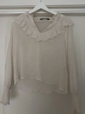 Vit blus med volang från Zara - Romantisk vit blus från Zara med diskret prickmönster, volangdetalj vid halsringningen och långa ärmar med smock vid ärmslut. Tillverkad i ett lätt och luftigt tyg som ger en feminin och trendig look.