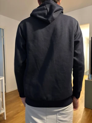 Mörkblå Champion hoodie med huva - Säljer en mörkblå hoodie från Champion med stor logga på bröstet och klassisk känguruficka. Hoodien har huva och ribbade muddar vid ärmar och midja. Perfekt för chill dagar eller när du vill ha en clean streetstyle-look.