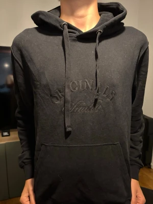 Svart hoodie med broderad text - Säljer en svart hoodie med broderad text 'ORIGINALS' på bröstet och stor magficka. Hoodien har dragsko i huvan och ribbade muddar vid ärmar och midja. Perfekt för chill dagar och enkel att matcha med jeans eller mjukis.