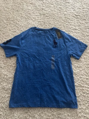 Blå t-shirt från Ralph Lauren - Snygg blå t-shirt från Ralph Lauren med klassisk logga broderad på bröstet och en siffra på ärmen. T-shirten har rund hals och är kortärmad, perfekt för en avslappnad stil. Tillverkad i mjuk bomull för extra komfort. Har storlek (14-16 L) vilket motsvarar S. Helt ny med prislapp.