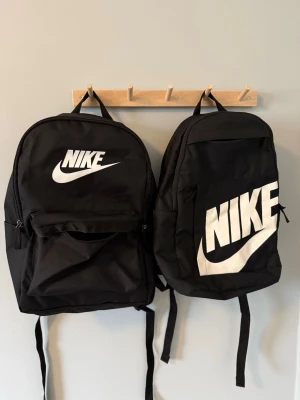 Svarta ryggsäckar från Nike - Två svarta ryggsäckar från Nike med stora vita logotyper på framsidan. Klassisk design med ett huvudfack, framficka med dragkedja och justerbara axelband. Tillverkade i slitstarkt syntetmaterial, perfekta för skola eller träning.