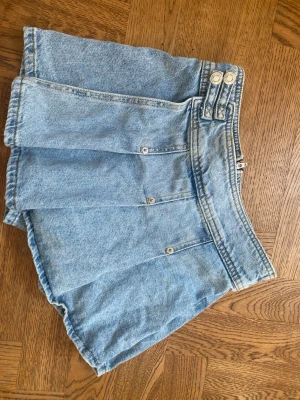 Kort ljusblå jeansskort - Säljer en kort ljusblå jeansskort med silvriga knappar och dragkedja framtill. Kjolen har markerade sömmar som ger en cool look. Perfekt för dig som gillar denim och vill ha en trendig stil.✨💕