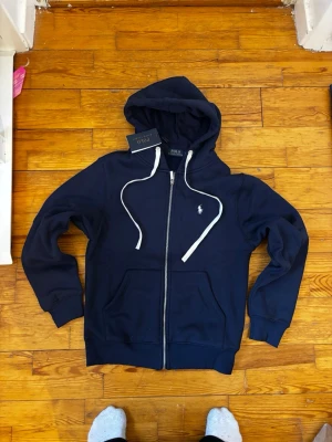 Mörkblå hoodie från Polo Ralph Lauren - Snygg mörkblå hoodie med dragkedja från Polo Ralph Lauren. Klassisk logga broderad i vitt på bröstet, vita dragsnören och två fickor framtill. Tillverkad i mjukt bomullsmaterial och har ribbade muddar vid ärmar och nederkant. Perfekt för en avslappnad stil.