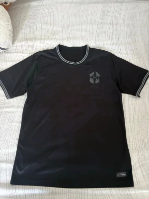 Svart Brasilien fotbollströja - Snygg svart fotbollströja med Brasilien-emblem på bröstet och fem stjärnor i nacken. Tröjan har korta ärmar, rund hals och vita detaljer på krage och ärmslut. Tillverkad i ett lätt och ventilerande material som passar perfekt till träning eller match.