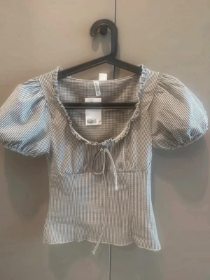 Randig puffärmad topp med snörning - Supersöt topp med smala svarta och vita ränder, puffärmar och snörning framtill. Toppen har croppad passform och en lätt rynkad kant vid urringningen. Perfekt för dig som gillar vintagevibbar och romantisk stil.