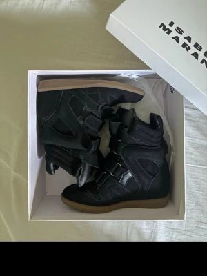 Svarta sneakers Isabel Marant - Säljer ett par svarta sneakers från Isabel Marant med högt skaft och kilklack. Skorna har en cool design med kardborreband och detaljer i mocka och läder. Yttersulan är i gummi och har en brun färg. Perfekta för dig som vill ha en trendig och unik look.