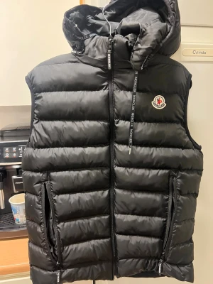 Svart dunväst från Moncler med huva - Snygg svart dunväst från Moncler med avtagbar huva och två dragkedjeförsedda fickor. Västen har Moncler-logga på bröstet och detaljerade dragkedjor med Moncler-tryck. Perfekt för lager-på-lager och streetwear-vibe.