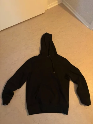 Svart basic hoodie med huva - En enkel svart hoodie med huva och dragsnören. Hoodien har långa ärmar, ribbade muddar vid ärmslut och nederkant samt en klassisk magficka framtill. Perfekt för en avslappnad stil och funkar året runt. Köpt på primark i Frankrike, enkel vardagshoddie som blivit för liten på mig 