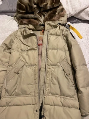 Beige pufferjacka med fuskpälsluva - Säljer en beige parajumper med stor luva fodrad i mjuk päls. Jackan har två stora fickor framtill med knappstängning och dragkedja. Detaljer som gul rem på axeln och quiltad design. Perfekt för kalla vinterdagar.