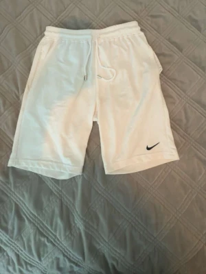 Vita shorts från Nike med snörning - Snygga vita shorts från Nike med elastisk midja och snörning framtill. Klassisk Nike-logga i svart på ena benet. Mjukt material som känns skönt mot huden och två sidofickor för det viktigaste. Perfekta för sommarens alla äventyr.