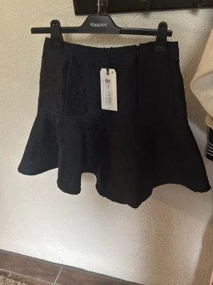 Svart volangkjol med struktur - Snygg svart kjol med volangkant och lätt utsvängd form. Kjolen har en diskret struktur i tyget och dragkedja i sidan för enkel påklädning. Perfekt för dig som vill ha en stilren men ändå lekfull look.