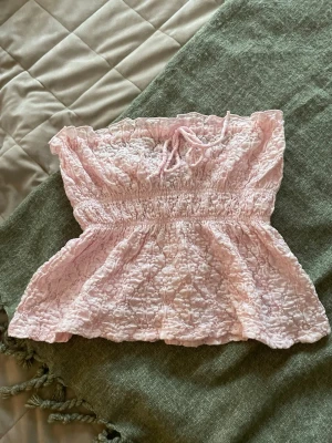 Ljusrosa spets tubtopp Gina Tricot - Supersöt ljusrosa tubtopp från Gina Tricot i spets med smockad midja och volangkant upptill. Toppen har en liten knytning framtill och är perfekt för sommarens alla outfits. Passar dig som gillar romantisk och feminin stil.