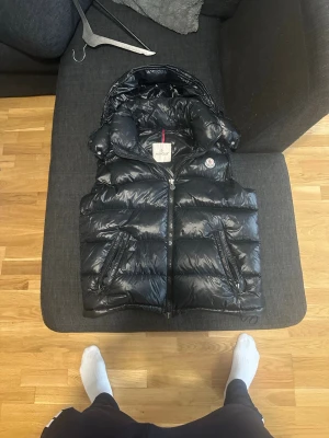 Svart dunväst från Moncler - Snygg svart dunväst från Moncler med glansig finish och avtagbar huva. Västen har två dragkedjeförsedda fickor och klassisk Moncler-logga på bröstet. Perfekt för lager-på-lager och håller dig varm under kyliga dagar.