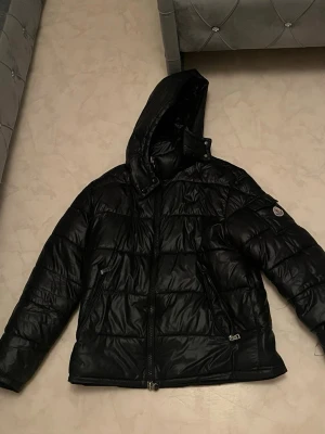 Moncler Maja  - Snygg svart pufferjacka från Moncler med glansig finish och vadderad design. Jackan har en stor huva, dragkedja framtill och två fickor med dragkedja. Moncler-logga på vänster ärm ger en exklusiv touch. Perfekt för kalla vinterdagar.