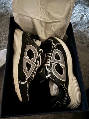 Svarta sneakers med vita detaljer - Säljer ett par svarta sneakers med vita och grå detaljer samt vita snören. Skorna har en chunky sula och stora CD-bokstäver på sidan. Materialet är en mix av mesh och syntet, vilket ger en sportig och modern look.