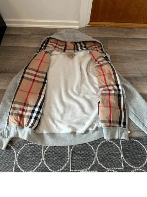 Grå hoodie med klassiskt rutigt foder - Snygg grå zip-hoodie med huva och dragsko. Insidan av huvan och öppningen har ett klassiskt beige, svart och rött rutigt foder som ger en exklusiv känsla. Två fickor framtill och ribbade muddar vid ärmar och midja. Perfekt för en chill och stilren look.