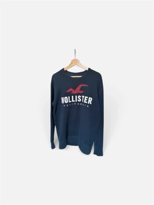 Mörkblå sweatshirt från Hollister - Mörkblå sweatshirt från Hollister med stor vit logga och röd fågel på bröstet. Klassisk rund halsringning och långa ärmar. Perfekt för en avslappnad och stilren look.