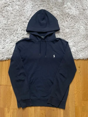 Mörkblå hoodie från Polo Ralph Lauren - Snygg mörkblå hoodie från Polo Ralph Lauren med klassisk huva och dragsnören. Har en vit broderad logga på bröstet och en stor magficka. Perfekt för en chill och stilren.  Står S men passar M. Som referens är modellen i bild 185cm