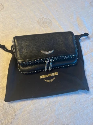 Svart kuvertväska från Zadig & Voltaire - Snygg svart kuvertväska i läder från Zadig & Voltaire med silverfärgade detaljer, dubbla dragkedjor och dekorativa nitar runt kanterna. Väskan har en ikonisk vinglogga framtill och levereras med en svart dustbag. Använts ett fåtal gånger så den är gott som ny💗