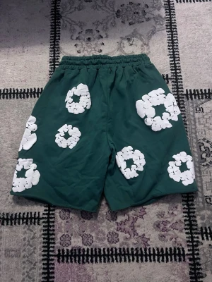 Gröna shorts med vita blommor - Mörkgröna shorts med elastisk midja och snörning. Shortsen har stora vita blommiga mönster tryckta över hela tyget och en avslappnad passform. Perfekta för sommarens alla äventyr.