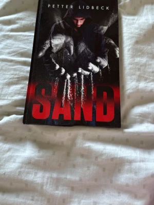 Sand av Petter Lidbeck - En spännande roman av Petter Lidbeck med svart och röd omslagsdesign. Boken handlar om Amad som får praktik på ett företag som jobbar med sand, där mystiska händelser förändrar hans liv. Perfekt för dig som gillar mysterier och ungdomsdrama.