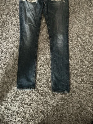 Nudie Mörkblå slitna jeans med hål - Säljer ett par mörkblå jeans med slitningar och stora hål vid knäna. Jeansen har en rak passform och klassisk femficksdesign. Perfekta för dig som gillar en edgy och avslappnad stil.