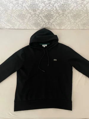 Svart hoodie från Lacoste - Säljer en svart hoodie från Lacoste med klassisk krokodil-logga på bröstet. Hoodien har dragsko i huvan och en stor magficka. Tillverkad i mjukt material som känns skönt mot huden. Perfekt för dig som gillar stilrena och tidlösa plagg.