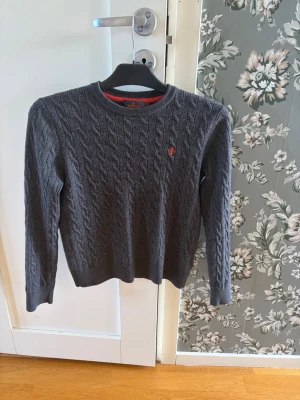 Mörkgrå kabelstickad tröja - Snygg mörkgrå kabelstickad tröja med rund hals och lång ärm. Tröjan har ett diskret rött broderat emblem på bröstet och ribbade muddar vid ärmslut och nederkant. Perfekt för lager på lager och passar till det mesta. Tröjan krympte i tvätten så kanske passar xs-xxs. Priset är ej hugget i sten. 