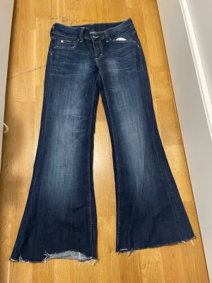 Mörkblå bootcut jeans med rå kant - Säljer ett par mörkblå bootcut jeans med slitningar och råa, fransiga benslut. Jeansen har klassisk femficksmodell och lätt tvättade detaljer framtill. Perfekta för dig som gillar en avslappnad och trendig look.