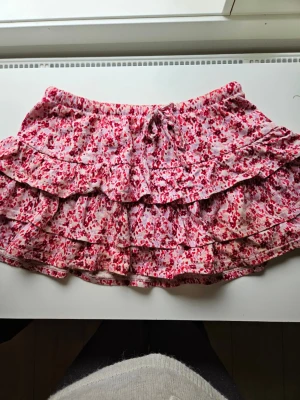 Volangkjol - Söt kort kjol med blommigt mönster i rosa och rött. Kjolen har flera volanger och ett knytband i midjan för extra detalj. Perfekt för vår och sommar när du vill ha en lekfull och luftig stil.