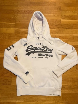 Vit Superdry hoodie Storlek S - Vit hoodie från Superdry med stort svart tryck på bröstet och japanska tecken. Klassisk känguruficka framtill, huva med dragsko och Superdry-detalj på snöret. Snygg och sportig stil och passar riktigt bra till nästan allt. Lite nopprig och trycket har blivit slitet.