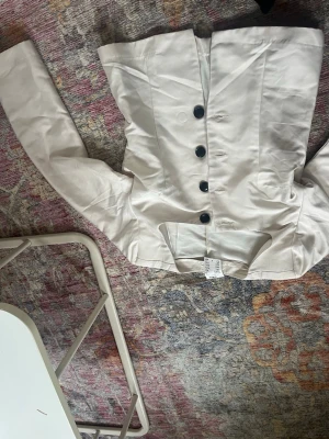 Vit kavaj med svarta knappar - Stilren vit kavaj med långa ärmar och fyra svarta knappar framtill. Kavajen har en rak passform och markerad krage, perfekt för en trendig och clean look. Materialet är slätt och ger en elegant känsla.