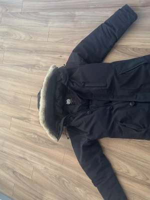 Svart canada goose parkas med riktig päls - Snygg svart parkas med huva och riktig päls. Jackan har dragkedja och knappar framtill, samt ribbade muddar vid ärmsluten för extra värme. Praktiska fickor på sidorna och klassisk längre modell som passar perfekt till kalla dagar. Jackan är i perfekt tillstånd förutom i ärmarna, det är den ända synliga skadan. storlek xs passar perfekt till 170-185cm 