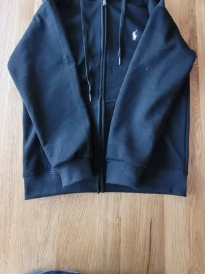 Svart Raulph Lauren kofta  - Snygg svart hoodie med dragkedja. Klassisk design med huva och dragsnören samt den ikoniska vita broderade loggan på bröstet. Tillverkad i mjukt bomullsmaterial och har ribbade muddar vid ärmslut och nederkant.