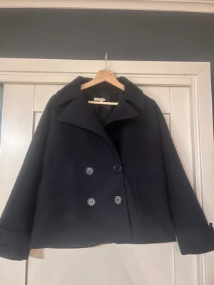 Mörkblå skepparkavaj med dubbelknäppning - Jag säljer min kappa som knappt är använd. Den är i gott skick och säljer den pågrund av att den ej kommit till användning. Pågrund av materialet så är den ganska nopprig.