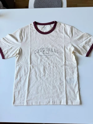Vit t-shirt med vinröda kanter - Vit t-shirt från Jack & Jones Originals med vinröda muddar vid hals och ärmslut. Tryck med texten 'ORIGINALS' på bröstet. Klassisk passform och tillverkad i mjuk bomull.