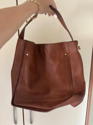 Brun bucket bag i läder - Snygg brun bucket bag i äkta läder med bred axelrem och dekorativa sömmar. Väskan har magnetlås och innerfack med dragkedja. Perfekt för dig som gillar stilrena och rymliga handväskor med en klassisk look.