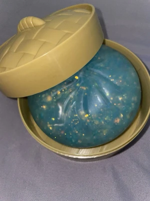 Blå squishy stressboll i plastkorg - En rund blå squishy stressboll fylld med glitter och färgprickar, formad som en bao och levereras i en beige plastkorg med lock. Bollen är gjord av mjuk plast och gummi, perfekt att klämma på när man vill slappna av eller bara leka.