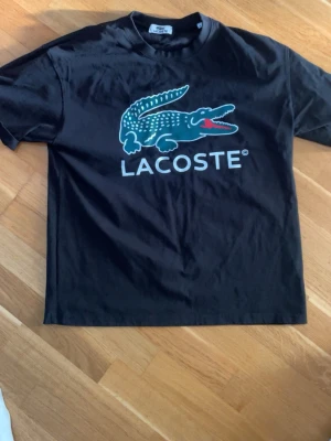 Svart Lacoste t-shirt med logga - Svart t-shirt från Lacoste med stor krokodil-logga och text på bröstet. Klassisk rund hals och korta ärmar. Tillverkad i mjuk bomull som är skön att bära. Perfekt för dig som gillar stilrena och sportiga plagg.