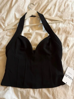 Svart halterneck korsettopp - Snygg svart korsettopp med halterneck och markerade kupor. Toppen har en croppad passform och är perfekt för att lyfta vilken outfit som helst. Tillverkad i ett slätt material som ger en stilren look.