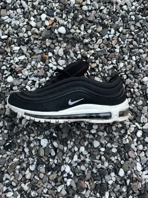Nike Air Max 97 svart/vit sneaker - Nike Air Max 97 i svart med vita detaljer och synlig Air-sula. Snygg vågig design på ovandelen och liten vit Swoosh på sidan. Skon har en låg silhuett och är tillverkad i en mix av syntet och textil.