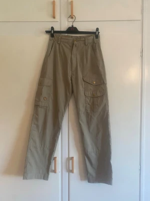 Fjällräven beiga byxor, - Säljer ett par beiga cargobyxor med flera praktiska fickor på sidorna och knappdetaljer. Byxorna har raka ben och bälteshällor i midjan. Perfekta för dig som gillar en avslappnad och funktionell stil. Materialet ser ut att vara slitstarkt bomullstyg.