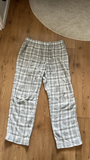 Gråvita rutiga pyjamasbyxor - Mysiga pyjamasbyxor i grått och vitt med klassiskt rutmönster. Byxorna har raka ben, resår i midjan och sidofickor. Perfekta för chillkvällar hemma eller när du vill ha det extra bekvämt.