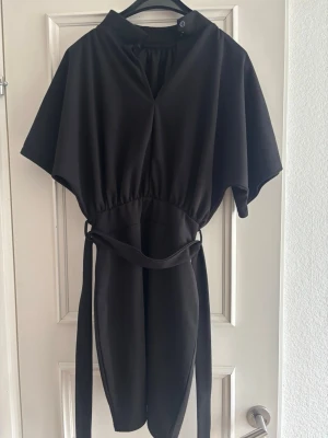 Svart kort klänning med bälte - Svart kort klänning med lös passform, breda korta ärmar och markerad midja med tillhörande bälte. Klänningen har en enkel, stilren design och knäppning vid halsen. Perfekt för dig som gillar minimalistisk stil.